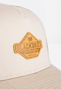 Beige cepure ar tekstūru audumu, ar brūnu ādas uzlīmi, uz kuras ir iegravēts "BLACKSKIES PREMIUM SUPPLIES MMIX".