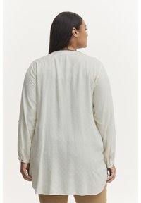 Blouse à manches longues de couleur beige clair avec un motif texturé et un ourlet arrondi. Possède des poignets à boutons et une coupe décontractée et modeste.