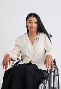 Blusa crema con maniche a sbuffo e chiusura con bottoni, abbinata a una gonna plissettata nera, indossata da una persona in sedia a rotelle.