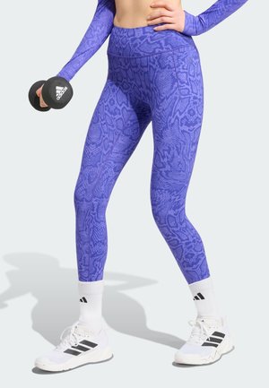 Person i lilla leggings med mønster og matchende top holder en sort Adidas håndvægt, iført hvide Adidas sneakers og sokker på lys baggrund.