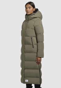 Lange olijfgroene pufferjas met een capuchon, voorzien van horizontale baffles, een ritssluiting zijzak en een label aan de zoom. Zachte, gladde textuur.