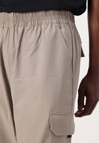 Lys beige cargo bukser laget av lett stoff. Har elastisk midje, sidelommer og en cargo-lomme med klaffdetalj.