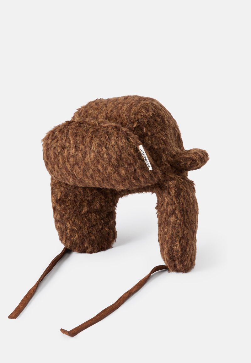 Barna szőrme sapka plüss textúrával, kerek tetejével és fülvédőkkel. Barna pántokkal rendelkezik. Oldalán egy kis márkajelzés található.