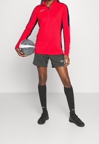 Punainen pitkähihaine Nike-paita, jossa on neljänneksen vetoketju ja mustat sivuetteet; Nike-logo rinnassa. Mustat kuviolliset shortsit, mustat polvisukat, mustat jalkapallokengät.