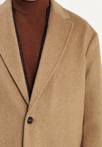 Cappotto di lana color cammello con un unico bottone nero indossato sopra un dolcevita marrone e con il colletto di una camicia a scacchi scura visibile sotto.