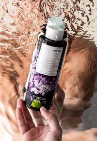 Flaska med lilafärgad kroppstvätt, svart cylindrisk design med blommönster, vit flip-top-kapp, 250 ml, placerad mot en reflekterande bakgrund.