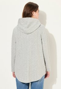 Žena s dlouhými hnědými vlasy, oblečená v lehké šedé prošívané bundě s kapucí a modrých džínách, stojící zády k jednoduchému světlému pozadí.