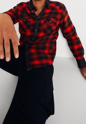 Hombre con una camisa de cuadros roja y negra abotonada y pantalones negros, con una mano apoyada en una superficie blanca y la otra extendida hacia adelante.