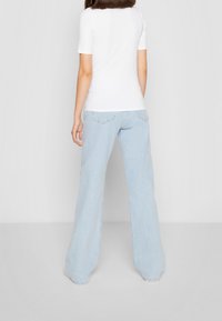 Žena stojící na bílém pozadí, oblečená v jednoduchém bílém tričku s krátkým rukávem a světlemodrých širokých kalhotách, odvrácená tváří.