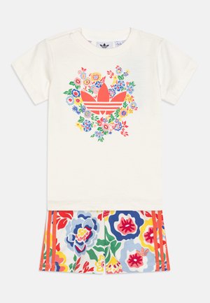 Kratkoroč T-shirt v kremni barvi z rožnatim grafičnim potiskom in rdečim logotipom, kombiniran z večbarvnimi kratkimi hlačami z rožnatim vzorcem in rdečimi črtami.