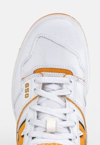 Białe sportowe sneakersy z pomarańczowymi akcentami, teksturowana skórzana cholewka, czarne detale, wyściełany język i płaskie białe sznurówki. Widać numer modelu 650.