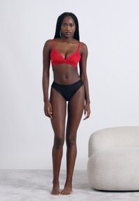 Piros melltartó és fekete bugyi; a fehérnemű puha anyagból készült, egyszerű kialakítású és vékony pántokkal rendelkezik. A modell világos, texturált felületen áll.