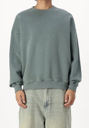 Homme portant un sweat-shirt à col rond ample de couleur verte sourde et un jean gris clair délavé, sur fond blanc.