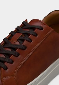 Baskets en cuir marron avec lacets noirs et semelle blanche, coutures détaillées le long du côté et œillets, vue en gros plan.