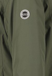 Giacca outdoor verde oliva con finitura testurizzata, dotata di una patch rotonda nera e argento del marchio sulla parte superiore del braccio e dettagli cuciti.