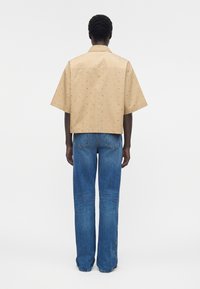 Persona de espaldas que lleva una camisa beige de manga corta con pequeños estampados y vaqueros azules de pierna recta sobre un fondo blanco.