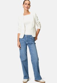 Blazer bianco su una maglietta bianca, abbinato a jeans a gamba larga blu chiaro. Presenta una chiusura con bottone e sneakers bianche casual.