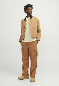 Chaqueta de mezclilla marrón claro con cuello negro, combinada con una camiseta verde claro y pantalones cargo en tono marrón claro, finalizada con botas beige. Diseño y textura simples.