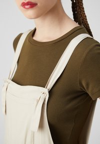 Beige overal s nastavitelnými popruhy, uvázanými na ramenou, nošený přes tmavě olivový žebrovaný trik. Hladká textilie, jednoduchý design.
