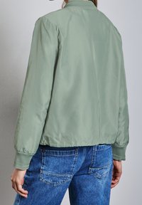 Světle zelená bomber bunda s hladkým povrchem, ribbovaným límcem a elastickými manžetami, v kombinaci s modrými džíny s vysokým pasem.