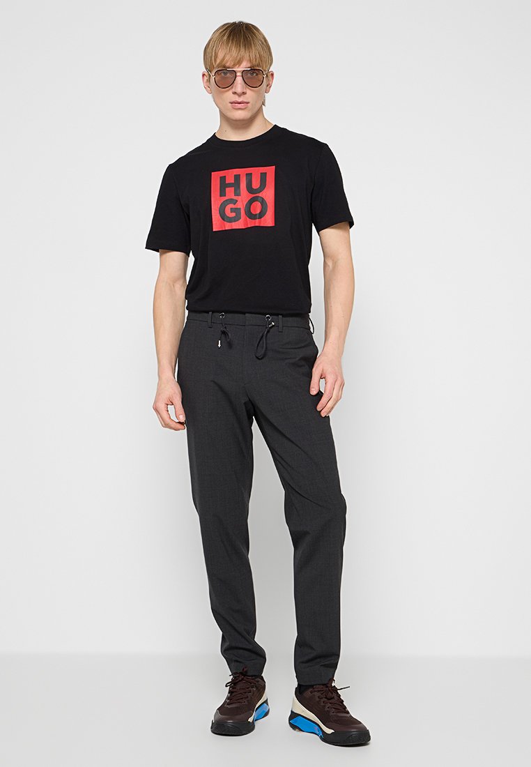 Schwarzes T-Shirt mit rotem "HUGO"-Logo, kombiniert mit dunkelgrauen, schmal zulaufenden Hosen mit Kordelzug und seitlichen Taschen, abgerundet mit braunen Sneakers.