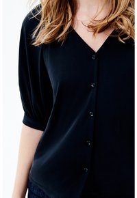 Blusa negra de manga corta con cuello en V, que presenta una textura suave y seis botones en la parte delantera. La tela tiene un ligero drapeado para un ajuste relajado.