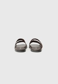 Des sandales en cuir marron présentent deux larges sangles croisant et une semelle plate. Le design est minimaliste avec une texture lisse et sans ornements.