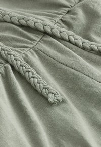 Tejido verde claro con un acabado texturizado, que presenta un detalle trenzado a lo largo de una costura, mostrando un suave caimiento y una sutil costura.