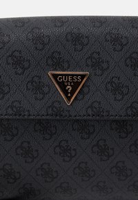 Černá texturovaná kabelka s jemným raženým vzorem a trojúhelníkovou kovovou plaketou s nápisem „GUESS USA“. Hladký povrch na okrajích.