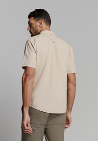 Korte mouw beige shirt van zacht materiaal, met een standaard kraag en een enkele knoopdetail achter. Getailleerde pasvorm, subtiele textuur.