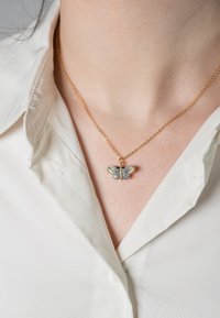 Collana in oro con ciondolo a forma di farfalla, con dettagli blu e verdi, indossata sopra una camicia bianca con colletto.