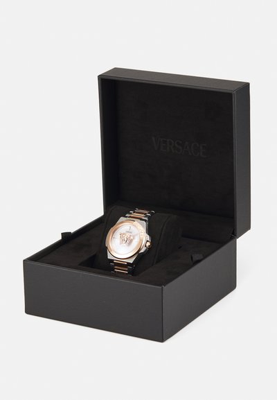 Versace laikrodis, pateiktas juodame dėkle. Jame yra rožinio aukso rėmelis, sidabro ir juodos spalvos juosta bei balta ciferblatas su liūto motyvu.