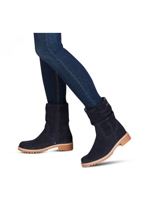 Personne portant des bottines en daim bleu foncé avec des semelles en caoutchouc marron clair et un jean skinny foncé, un pied légèrement levé.