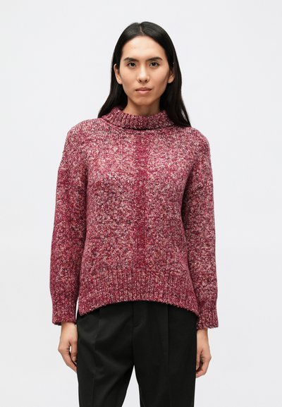 Kvinde med langt mørkt hår iført en rød og pink meleret strikket sweater og sorte bukser, stående mod en ensfarvet lys baggrund.