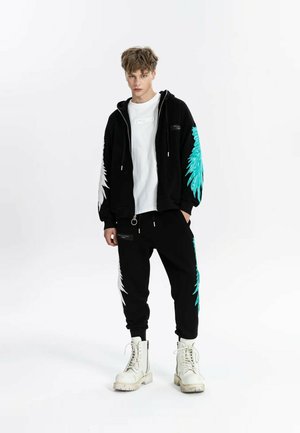 Joven está de pie, vistiendo una sudadera con capucha negra y pantalones con diseños de alas blancas y turquesas, camiseta blanca y botas blancas con cordones.