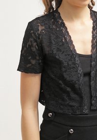 Černý krajkový bolero s krátkými rukávy, zdobený vlnitými okraji a květinovým vzorem. Nositeli přes černý top na ramínka s texturovanou látkou.