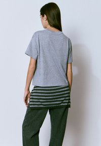 Camiseta de algodón gris con mangas cortas, que presenta un dobladillo en capas con rayas horizontales en negro y gris en la parte inferior, combinada con pantalones oscuros.