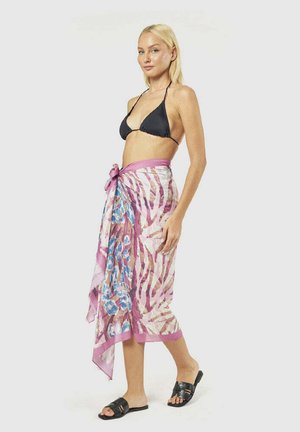 Donna bionda che indossa un top bikini nero, un sarong rosa e decorato legato in vita e sandali neri scivolati, in piedi di lato.