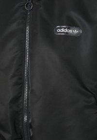 Manteau rembourré noir avec une fermeture éclair sur le devant, présentant une finition lisse et un patch transparent du logo adidas sur le côté gauche.