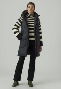 Črna podložena usnjena brezrokavnik s kapuco, oblečen nad črtasto črno-belo puloverjo. Hlače so črne, obutev pa so zelene škornje s peto.