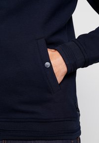 Handen inskjuten i framfickan på en mörk marinblå strukturerad tröja med synliga detaljer på ärmsluten och en märkesknapp.