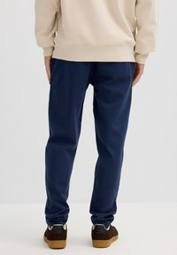 Persona con pantalones azul marino, sudadera beige y zapatillas marrones de pie sobre un fondo blanco liso, de espaldas.