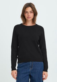Zwarte gebreide trui met lange mouwen, ronde hals en geribde zoom in een relaxte pasvorm. Gecombineerd met denim jeans, tegen een lichte achtergrond.