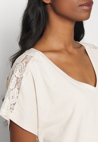 Béžové tričko s výstřihem do V má krajkovou aplikaci na ramenech, měkkou texturu a volný střih.