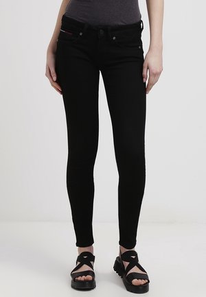 Kobieta ubrana w czarne obcisłe dżinsy i czarne sandały na platformie, stojąca na tle jasnego, jednolitego tła.