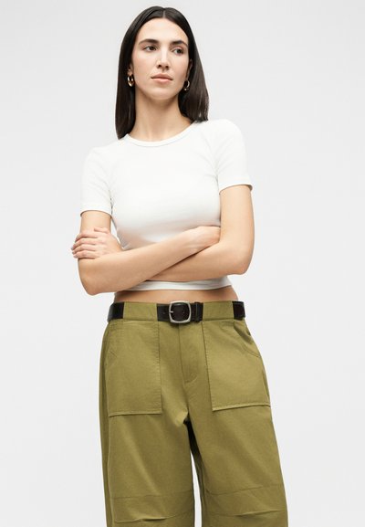 Γυναίκα με ίσια σκούρα μαλλιά που φοράει χρυσούς κρίκους σκουλαρίκια, λευκό κοντομάνικο crop top και παντελόνι ψηλόμεσο σε ελιά πράσινο χρώμα με μαύρη ζώνη.