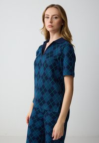 Polo a maniche corte di colore blu scuro con un motivo a rombi in teal e blu navy, caratterizzato da un colletto a V e un colletto blu navy uniforme.