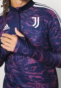 Pitkähihainen vetoketjullinen takki laivastonsinisellä pohjalla, jossa on vaaleanpunainen abstrakti kuvio. Takissa korkea kaulus ja peukaloreiät. Mukana Adidas- ja Juventuksen logot.