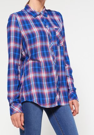 Kvinde iført en blå, rød og hvid ternet skjorte med lange ærmer og blå jeans, stående foran en ensfarvet hvid baggrund.