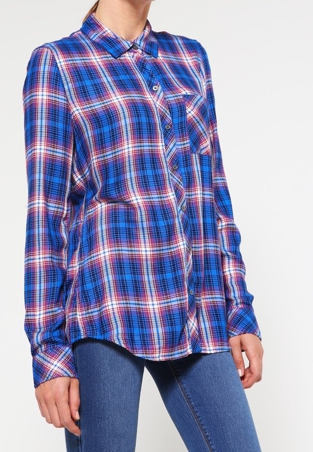 Femme portant une chemise à carreaux bleue, rouge et blanche à manches longues et un jean bleu, debout devant un fond blanc uni.